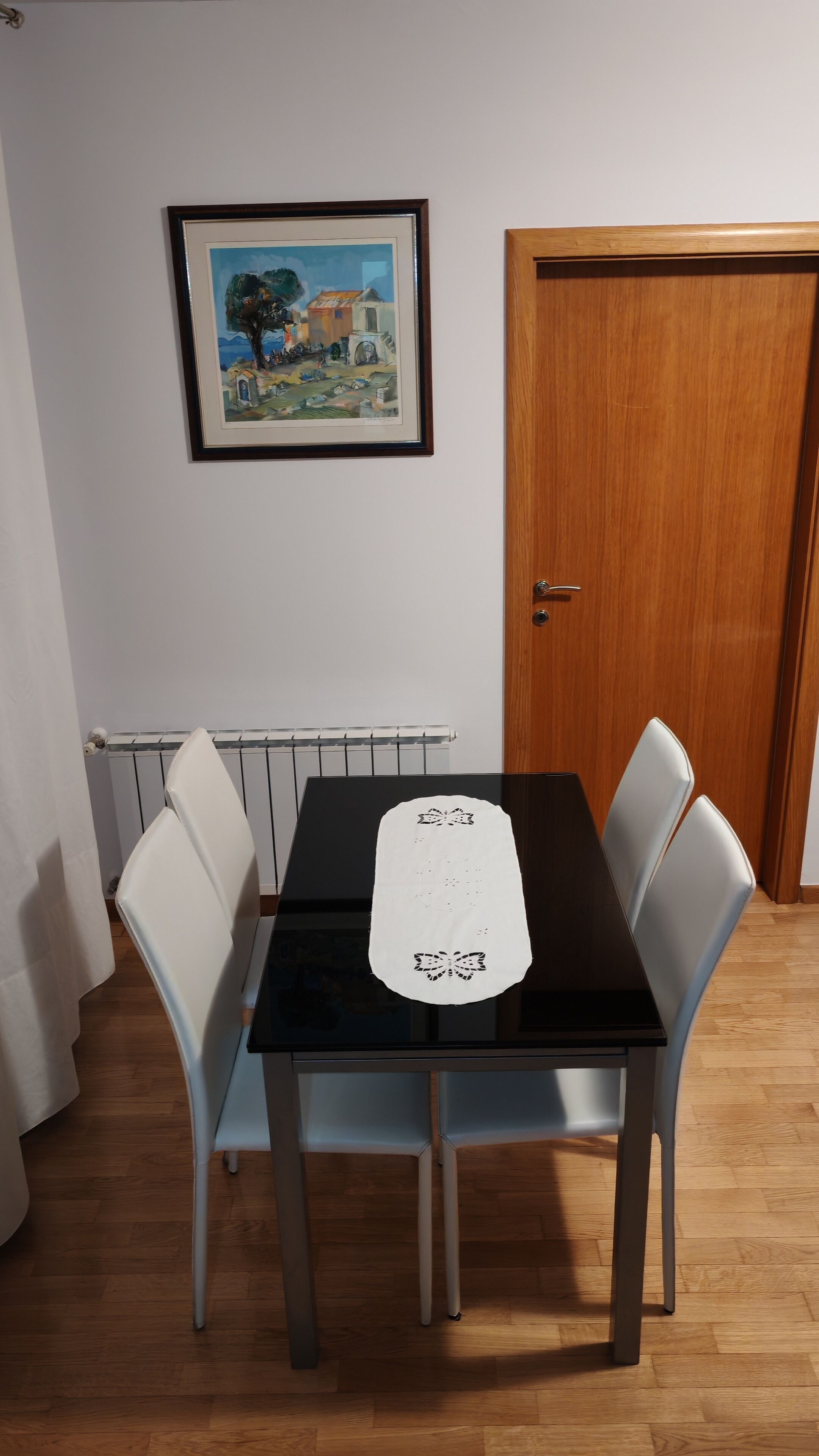 Ein Esstisch mit vier Stühlen, ein gerahmtes Bild an der Wand, ein Heizkörper darunter und eine Holztür zur rechten Seite.