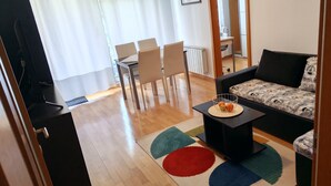 Una sala de estar con un sofá, una mesa de centro con fruta, un comedor con sillas y una cocina al fondo.