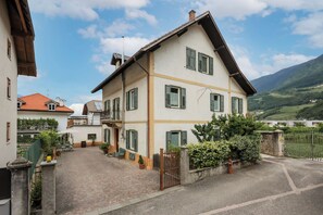 Une maison à deux étages avec une allée pavée, une entrée fermée par un portail et une vue sur une vallée verdoyante.