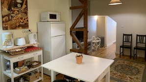 Une cuisine compacte avec un réfrigérateur blanc, un four à micro-ondes et une machine à café posés sur le plan de travail. On y trouve une table blanche ornée d’une plante en pot, ainsi qu’une chaise en bois dans l’espace repas.