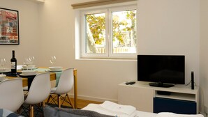 Ein Zimmer mit Esstisch für zwei Personen, einem Fernsehgerät auf einem Schrank und einem Fenster mit Blick auf Bäume.