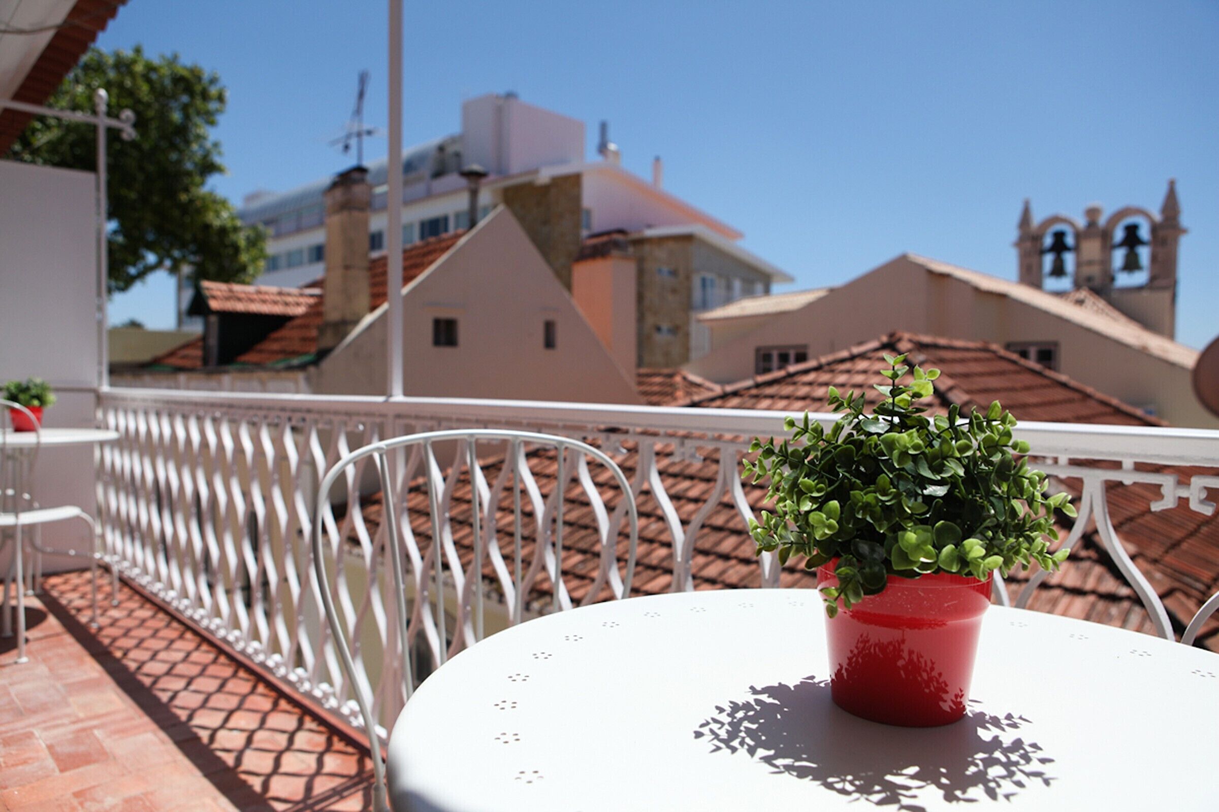 A balcony with a white table and chairs, a red potted plant, and a view of buildings with red roofs and a church tower.