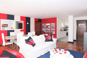 Ein modernes Wohnzimmer mit einem weißen Sofa, roten und schwarzen Kissen, einem Essbereich mit Holztisch und einer Küche im Hintergrund.