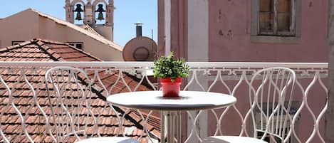 Un balcon avec une petite table ronde et deux chaises blanches, un pot de fleurs et une vue sur un bâtiment doté d’un clocher et d’une façade rose.