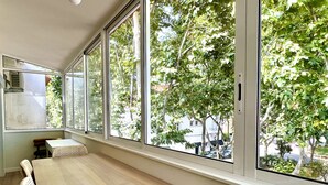 Un comedor con una larga mesa de madera y sillas de mimbre, ventanas amplias con vistas a árboles y edificios.