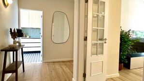 Una habitación con piso de madera, una puerta blanca con paneles de vidrio, un espejo ovalado instalado en la pared, una pequeña mesa de madera con un tazón y elementos decorativos, y vista a otra habitación con una encimera y alacenas.