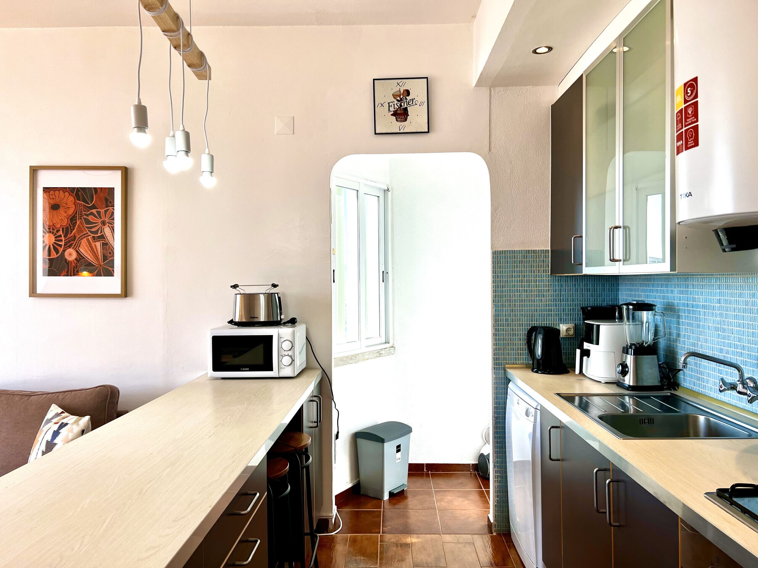 A modern kitchen with a microwave, toaster, and coffee maker. There is a sink, cabinets, and a trash bin. A framed picture hangs on the wall, and a window provides natural light.