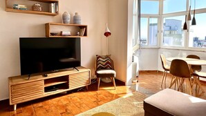 Ein modernes Wohnzimmer mit Fernseher, Holzregalen, einem Essbereich mit Tisch und Stühlen sowie einem großen Fenster mit Blick auf die Stadt.