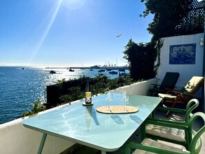 Eine Terrasse mit Tisch und Stühlen mit Blick auf das Meer, im Hintergrund Boote und eine Brücke.
