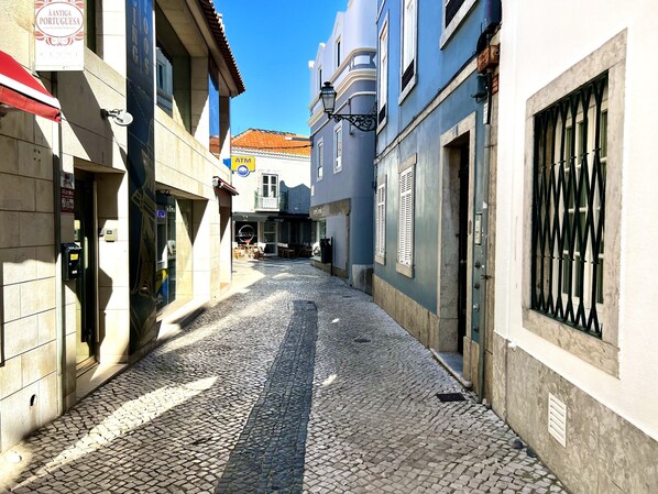 Eine enge Kopfsteinpflasterstraße mit Gebäuden an beiden Seiten, darunter ein sichtbarer Geldautomat und ein Schild für ein portugiesisches Restaurant.
