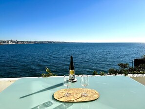 Ein Tisch mit einer Flasche und zwei Gläsern auf einer Matte, mit Blick auf ein Gewässer und eine ferne Stadtlandschaft.