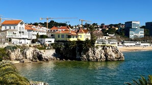 Un pueblo costero con edificios sobre un promontorio rocoso, una playa y grúas de construcción al fondo.