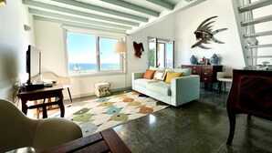Una sala de estar con sofá, una mesa de centro, televisor y vista al mar a través de una ventana.