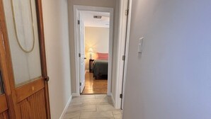 En hall med en trädörr till vänster, en lampa på höger sida och en säng med en röd sänggavel i bakgrunden.
