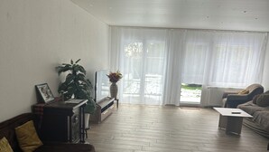 Ein geräumiges Wohnzimmer mit Holz-Fischgrätparkett, einem dunkelbraunen Sofa, einem weißen Sessel und einem kleinen Beistelltisch mit einer Blumenvase.