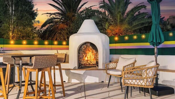 Een terras met rieten stoelen en een tafel, een ingebouwde open haard, en een decoratieve muur met lichtjes.