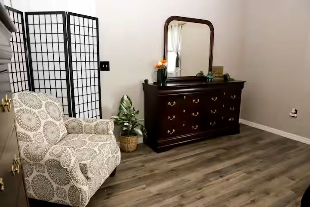 Une pièce avec un fauteuil à motifs, une commode en bois foncé dotée d’un miroir et un plancher en bois.