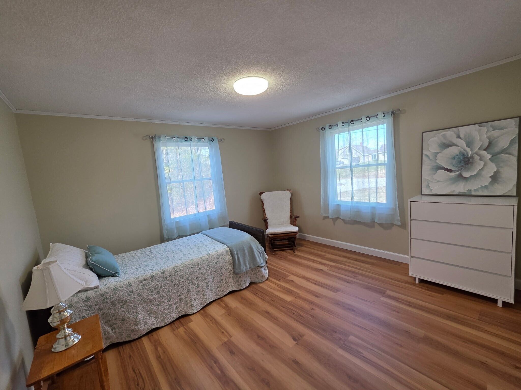 Un dormitorio con cama, silla, cómoda y dos ventanas con cortinas.