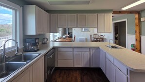 Una cocina con armarios blancos, un lavamanos de acero inoxidable, lavavajillas y un comedor con sillas de madera y una mesa.