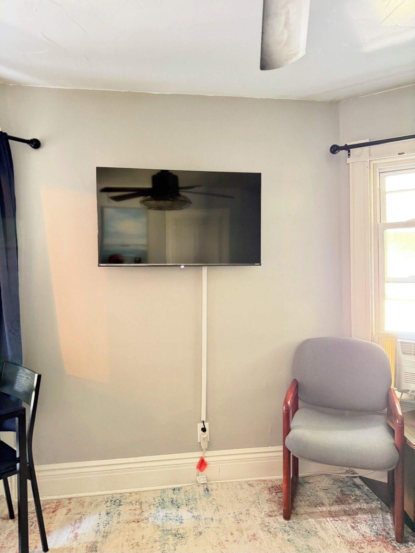 A room with a mounted flat-screen TV, a ceiling fan, a chair, and a window with curtains.