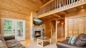 A wooden cabin interior with a stone fireplace, a television, a staircase, and a large wooden door. - Amazing Grace - Charming Cabin (Sevierville)