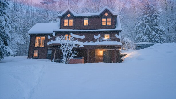 Ein großes, zweistöckiges Haus mit verschneitem Dach und Balkon, umgeben von schneebedeckten Bäumen.