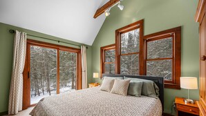 Ein Schlafzimmer mit einem großen Bett, zwei Nachttischen und Blick auf einen verschneiten Wald durch große Fenster.