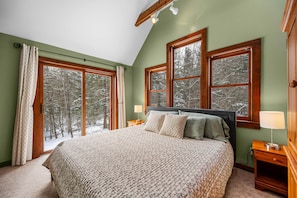 A bedroom with a large bed, two bedside tables, and a view of a snowy forest through large windows. - Stowe Hollow Retreat. Hot Tub, Pet Friendly (Stowe)