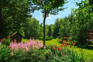 A lush garden with a wooden cabin, dense trees, and a variety of flowers. - Stowe Hollow Retreat. Hot Tub, Pet Friendly (Stowe)