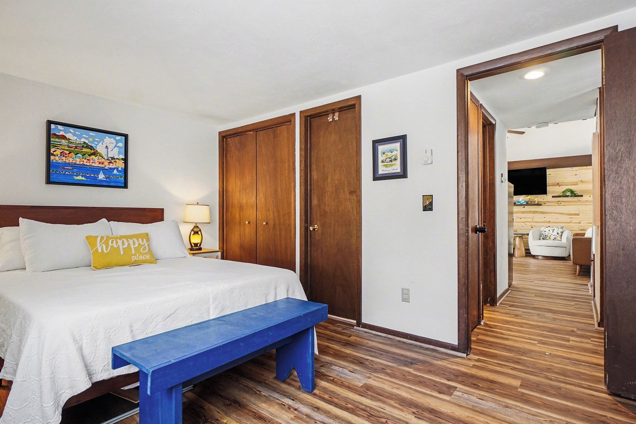 A hotel room with a bed, a bench, wooden flooring, a painting, and a wooden wardrobe.