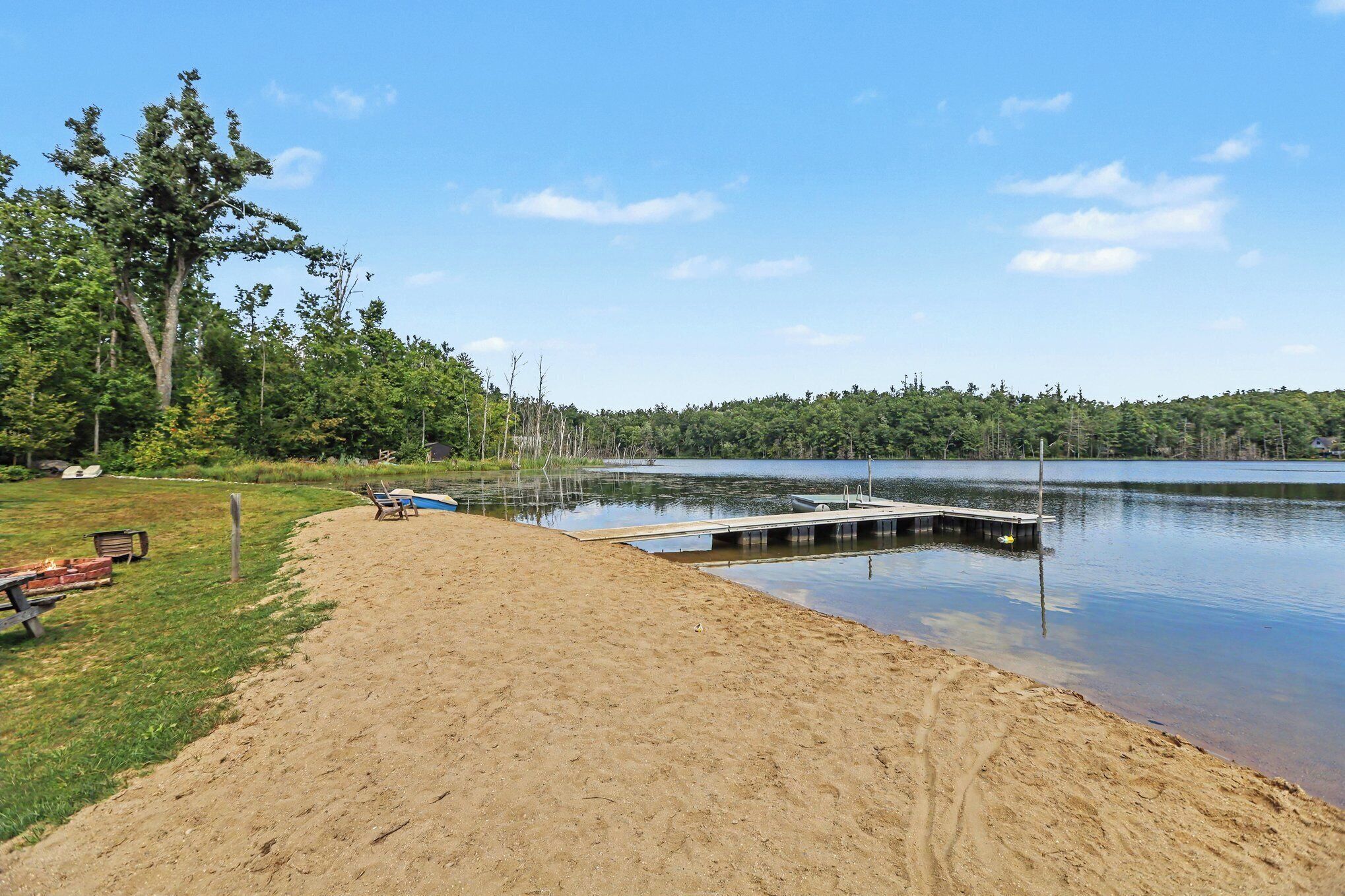 A sandy beach with a dock extending into a calm lake, surrounded by trees and a clear sky.