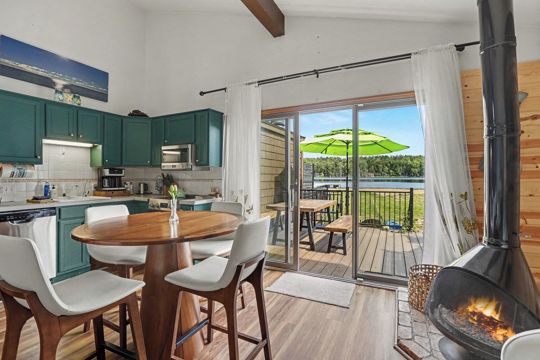 A modern kitchen with green cabinets, a dining area with a wooden table and chairs, and a large sliding glass door leading to a deck with a view of a lake.