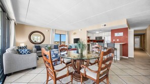 A modern living room with a glass dining table surrounded by wooden chairs, a grey sofa, and a large mirror on the wall. - Lucky 711 Island Winds East 3BD, 2BA Condo (Gulf Shores)