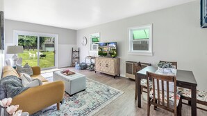 A modern living room with a sofa, ottoman, and dining table. There is a TV, a clock, and a window with blinds. - Downtown Perrysburg Bungalow w/ additional space (Perrysburg)