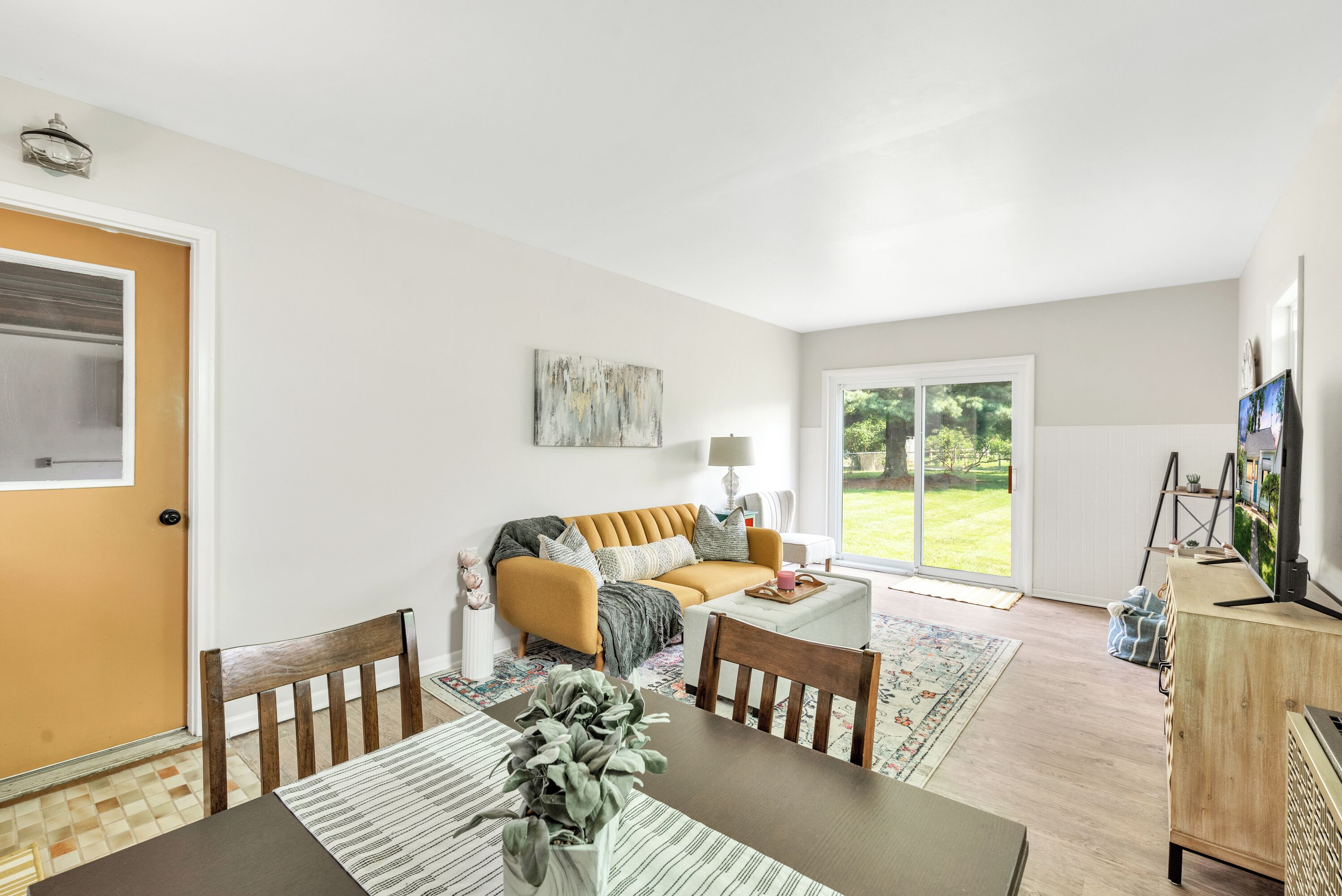 A modern living room with a sofa, coffee table, and dining table. There is a door with a window, a lamp, and a painting on the wall.