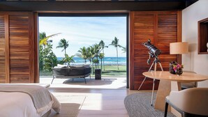 Un dormitorio con un ventanal grande que da a una playa, un telescopio sobre una mesa y una zona de estar acogedora.