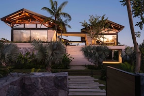 A modern house with a wooden deck, large windows, and a stone pathway leading to the entrance. - Modern Beachfront Villa: Chef, Pool & Premier Access (Punta Mita)