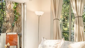 Un dormitorio con una cama grande, ropa de cama floral, una mesita de noche con lámpara y vistas a la vegetación por la ventana.