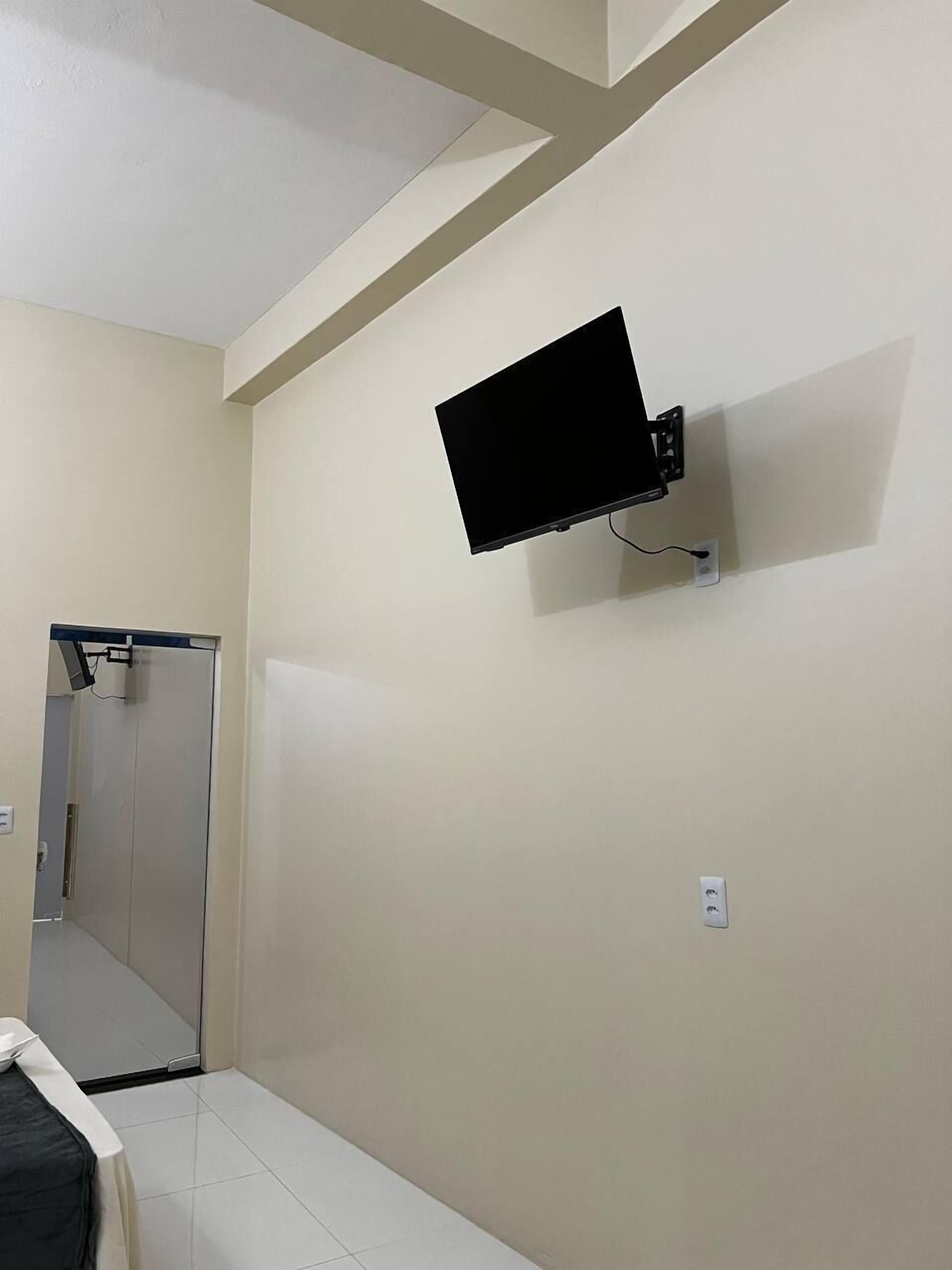 A room with a mounted flat-screen TV on the wall, a door leading to another room, and a bed with a white cover.