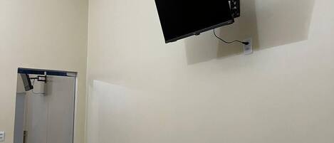 A room with a mounted flat-screen TV on the wall, a door leading to another room, and a bed with a white cover.