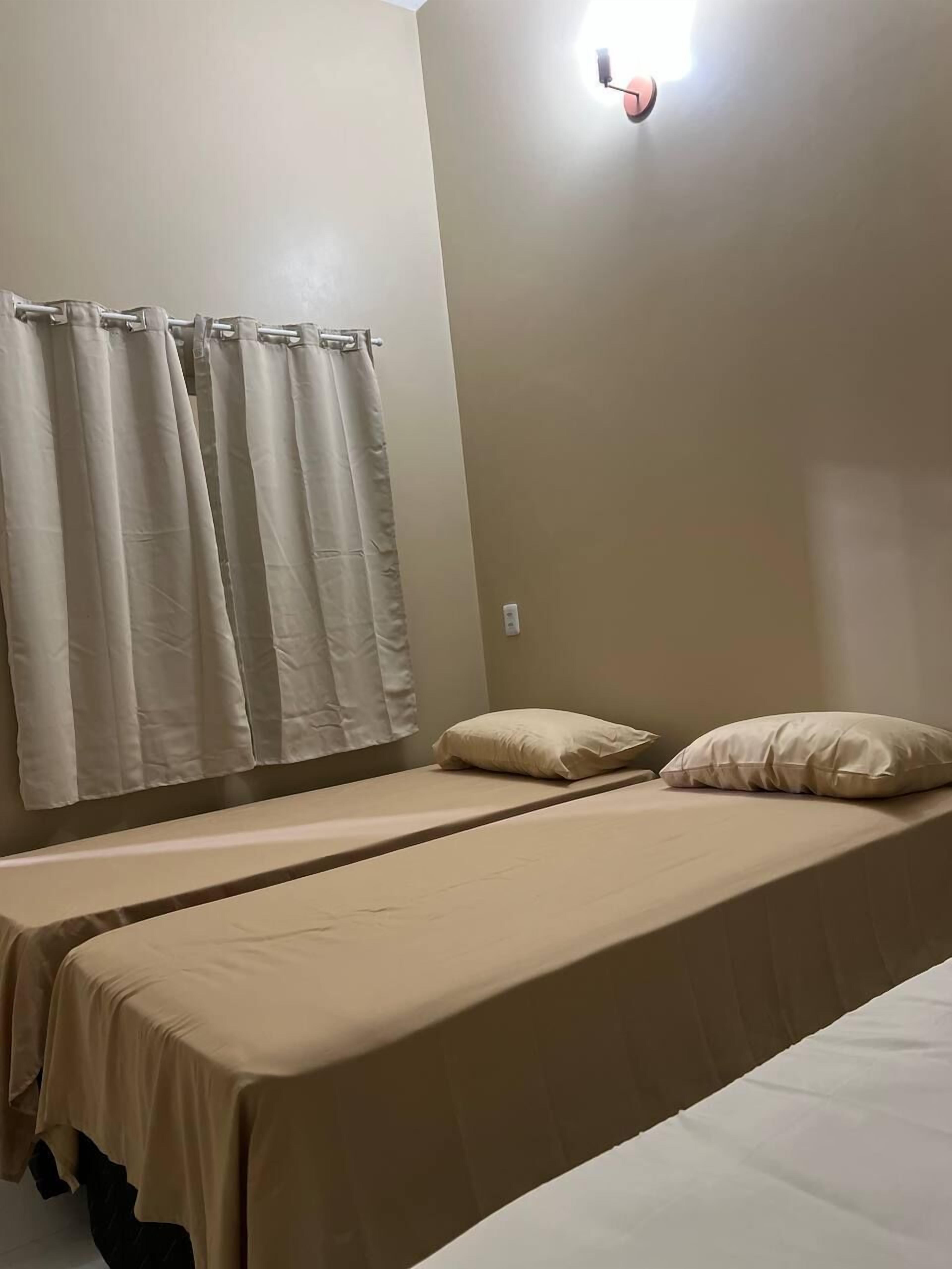 Un lit simple avec une couvre-lit beige et deux oreillers, une lampe fixée au mur et un rideau.