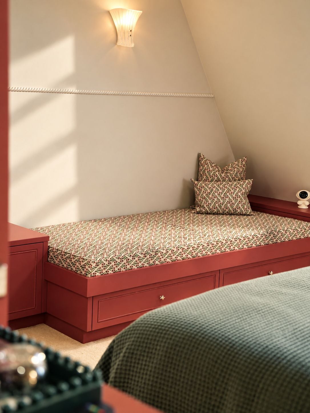 Una habitación pequeña y acogedora con techo inclinado, una cama con ropa de cama estampada, una mesita de noche con lámpara y una luz decorativa en la pared.