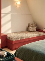 Ein kleiner, gemütlicher Raum mit schrägem Dach, einem Bett mit gemusterten Textilien, einem Nachttisch mit Lampe und einer dekorativen Wandleuchte.