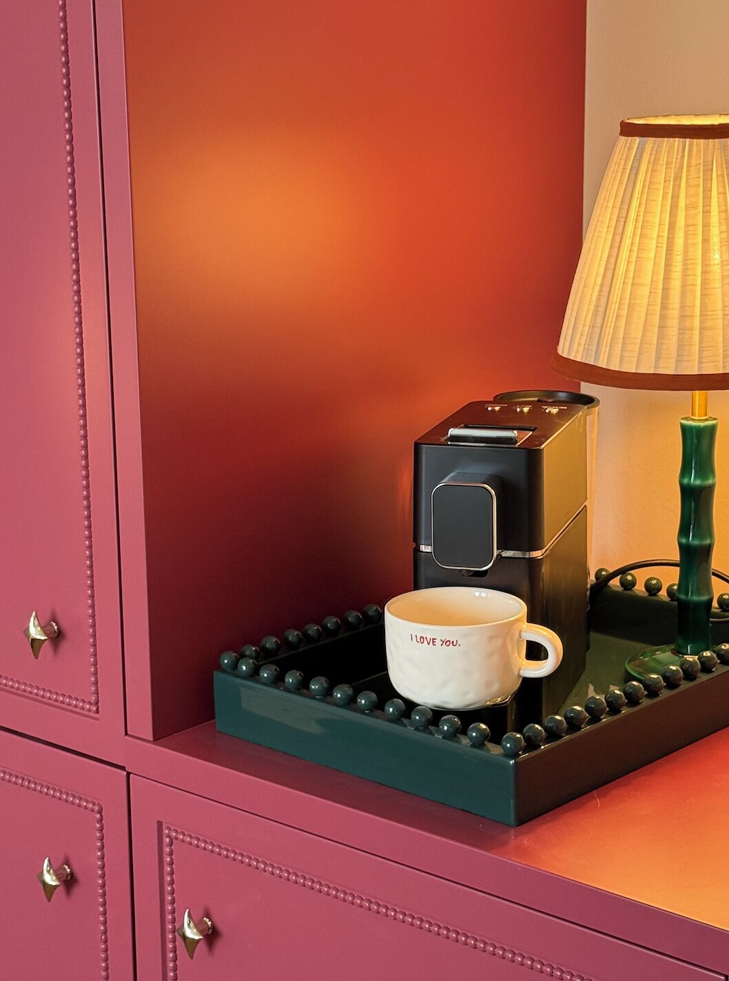 A coffee machine with a cup on a tray, a lamp, and a pink cabinet with drawers.