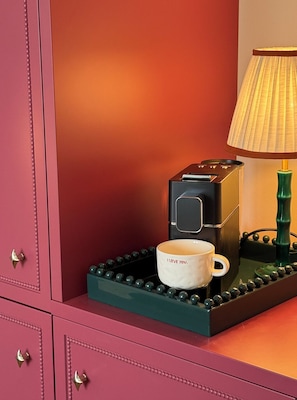 Eine Kaffeemaschine mit Tasse auf einem Tablett, eine Lampe und eine rosa Kommode mit Schubladen.