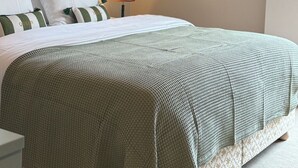 Un lit bien fait, recouvert d’une couvre-lit à motifs verts et blancs, avec une lampe de chevet et un tableau abstrait accroché au mur.