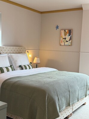Ein ordentlich gemachtes Bett mit einer Bettdecke in Grün-Weiß-Muster, einer Nachttischlampe und einem an der Wand hängenden abstrakten Gemälde.
