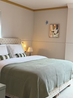 Ein ordentlich gemachtes Bett mit einer Bettdecke in Grün-Weiß-Muster, einer Nachttischlampe und einem an der Wand hängenden abstrakten Gemälde.