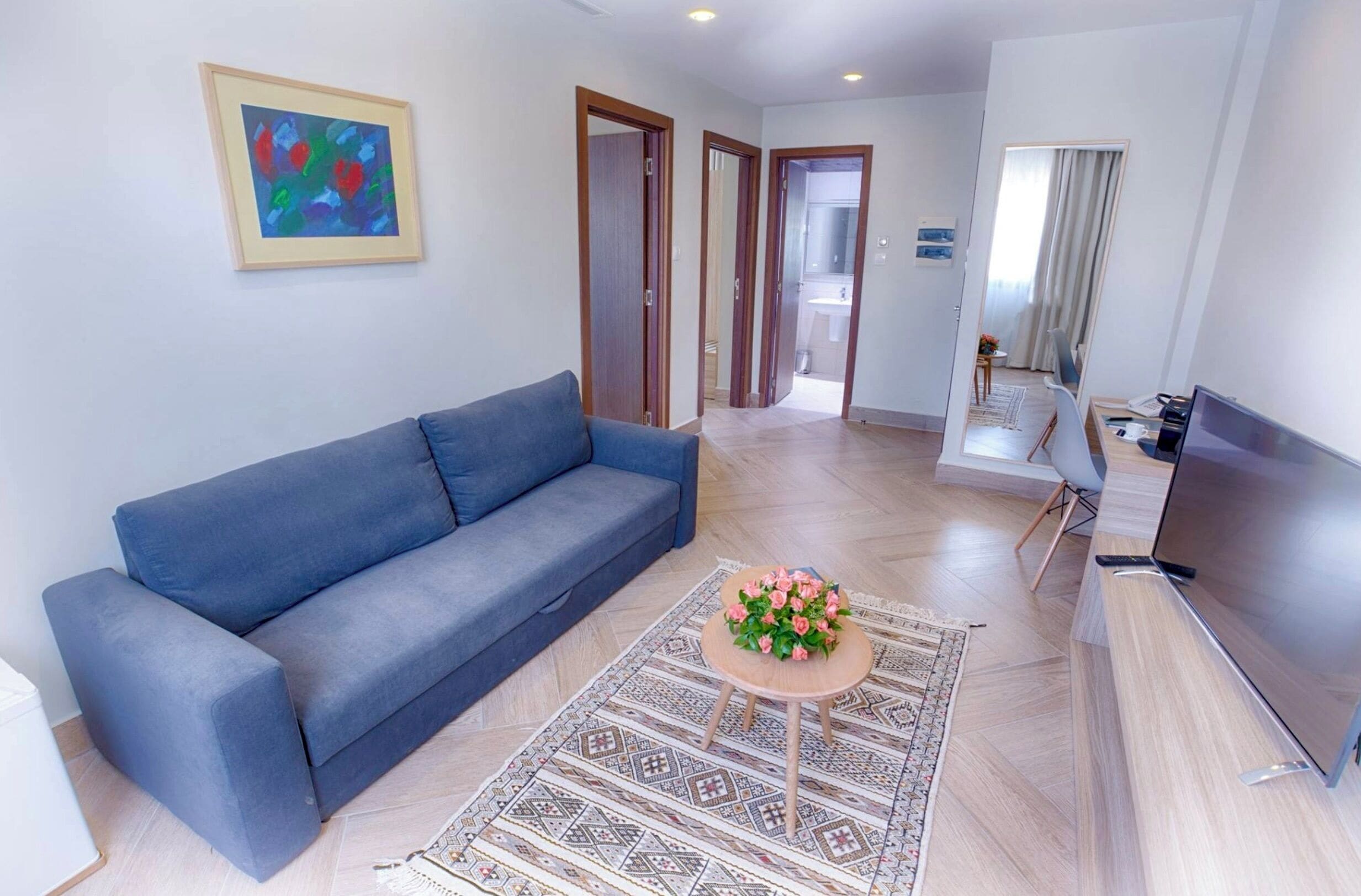 A modern living room with a blue sofa, a small round coffee table with flowers, a flat-screen TV, and a patterned rug.