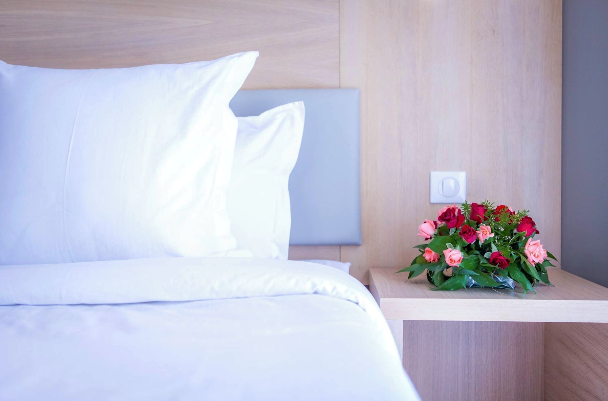 A neatly made bed with white linens and a wooden nightstand beside it. A bouquet of flowers is placed on the nightstand.